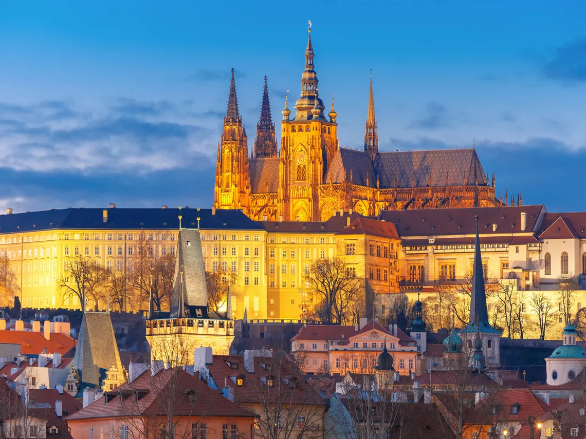 Večerní pohled na osvětlený Pražský hrad s dominantní Katedrálou svatého Víta a přilehlé historické budovy, obklopené městskou zástavbou v pozadí oblohy měnící barvy od modré k oranžové