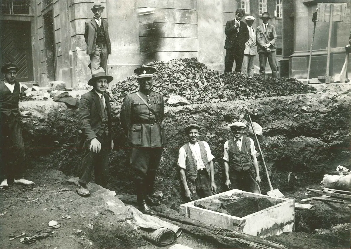 Historická černobílá fotografie ukazuje skupinu mužů, včetně vojáka a dělníků, na staveništi, obklopeni stavebním materiálem a vykopávkami, což naznačuje renovaci nebo výstavbu v minulém století